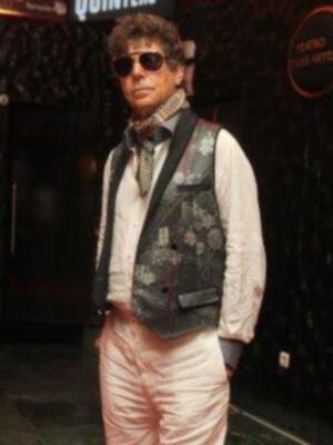 Un hombre con una chaqueta estampada y pantalones rosa claro, posando frente a un edificio con letras en el fondo.