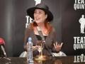 Una mujer con un sombrero negro habla en una conferencia de prensa, frente a un micrófono y una botella de agua. La imagen se centra en el Teatro Quintero, con un fondo que incluye su nombre y una ilustración de un personaje.
