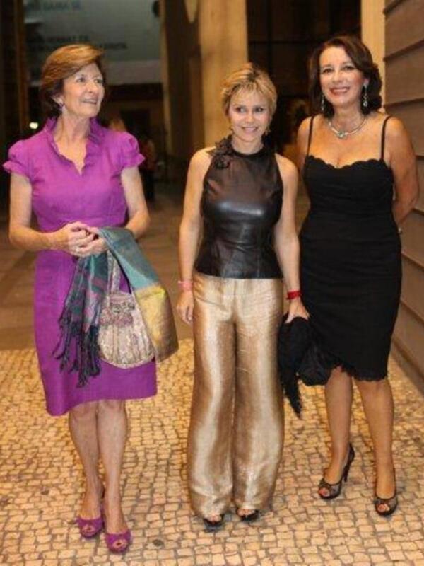 Tres mujeres posan juntas en un evento formal, dos de ellas con vestidos largos y una con un vestido negro. La imagen muestra detalles como joyas, bolsos y zapatos de tacón.