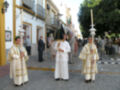 Imagen de una procesión religiosa con sacerdotes vestidos con hábitos blancos y dorados, portando candelabros. En el fondo, edificios con balcones y gente observando la procesión.