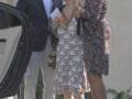 Una persona vestida con un traje azul y gafas de sol se dirige a una mujer en un vestido floral que sostiene un libro y una bolsa. Detrás de ellas, se puede ver la parte delantera de un automóvil blanco.