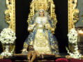 Virgen María en trono dorado, vestida con manto azul y blanca, rodeada de flores blancas.