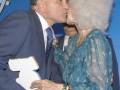 Una pareja en un evento formal, el hombre sostiene un trofeo mientras la mujer lo besa. El fondo es azul con letras blancas que dicen "VIII".