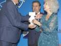 Un hombre en traje azul y una mujer en vestido verde están recibiendo un premio en un evento formal. El fondo es azul con una insignia y texto que parece ser un evento de premios o reconocimiento.