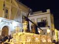 Un trono dorado con una figura vestida de negro y sosteniendo un crucifijo, frente a la Casa de la Cultura Alcalá. Banderas ondean en el cielo nocturno.