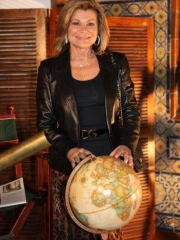Una mujer con cabello largo y dorado, vistiendo una chaqueta de cuero y un vestido negro, sostiene un globo terrestre. El fondo muestra detalles de una decoración con azulejos y madera.