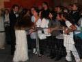 Una mujer con un abrigo de piel marrón y una falda larga blanca se acerca a un grupo de personas detrás de una barrera metálica. La escena parece ser en un evento formal, con la mayoría de las personas vestidas formalmente.