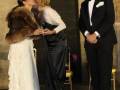 Una mujer en un vestido blanco con volantes se besa con otra mujer en un vestido negro, mientras un hombre en traje negro observa.