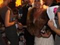 Una celebridad en un evento nocturno, posando con una peluca de piel y un vestido blanco. Algunos asistentes la rodean, uno con una micrófono y otro con un papel. La escena tiene luces de fondo que sugieren una celebración o premiación en un ambiente urbano.