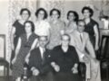 Una familia reunida en una foto antigua, con miembros de generaciones anteriores sentados y otros posando para la cámara. La escena muestra una atmósfera familiar y afectiva, con detalles de vestimenta y decoración típicos del siglo XX.