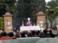 Una imagen de una ceremonia religiosa con un sacerdote en el centro, frente a una tribuna cubierta de blanco. En el fondo, se ven estatuas y un muro con una cruz. La audiencia está sentada en bancos, observando la ceremonia.
