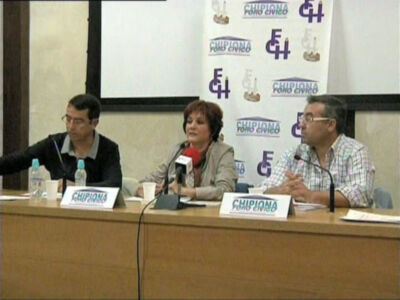 Panel de debate en el Foro Cívico de Chipiona, con tres participantes en una mesa con micrófonos y carteles con el logo del evento.