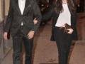 Un hombre y una mujer caminan juntos por la calle. El hombre lleva un traje negro formal, mientras que la mujer usa una chaqueta larga negra y pantalones ajustados con un bolso de mano. La imagen parece ser de una celebración o evento formal, dado el estilo de vestimenta y la presencia de otros individuos en el fondo.