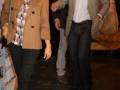 Una pareja caminando juntos en una alfombra roja. La mujer lleva una chaqueta beige y botas marrones, mientras que el hombre está vestido con una chaqueta beige y una camisa blanca. El fondo es oscuro, destacando su figura.