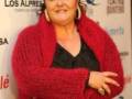 Una mujer con cabello rojo corto y ojos oscuros, vistiendo una chaqueta roja con un cuello alto y mangas largas. Su expresión es amigable, con una sonrisa ligeramente curvada y los ojos abiertos. La imagen está en un contexto de evento, con una bandera o cartel de fondo que contiene texto.