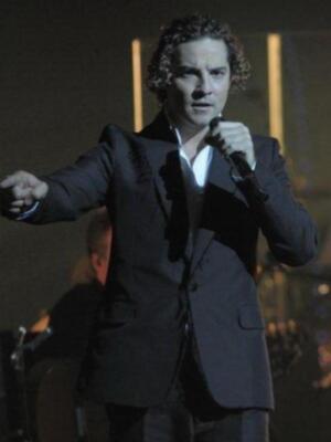 Artista cantando en escenario, vestido con traje negro y sosteniendo un micrófono.