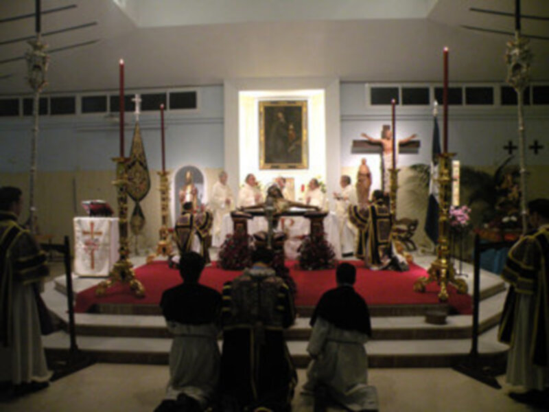 Imagen de una iglesia con un altar central, sacerdotes vestidos en trajes blancos y negros, una cruz grande en el centro, velas encendidas, un reloj de mano y un cuadro en la pared.
