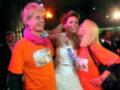 Una mujer con un vestido blanco y una corona de Miss Mundo, abrazada por dos personas en camisetas naranjas.