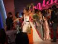 A una mujer vestida con un traje blanco y rojo brillante, posiblemente en un evento de belleza o concursos. Ella sostiene una corona y está rodeada por otros participantes vestidos de manera similar, con teléfonos y cámaras en sus manos. El fondo es rojo con letras blancas que parecen decir "Miss Universe".