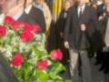 Una imagen de una procesión religiosa con personas vestidas formalmente, algunos portando flores rojas y blancas. La escena se desarrolla en un entorno urbano con edificios de fondo y una calle pavimentada.
