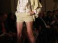 Una modelo camina por la pasarela de una feria de moda, luciendo un conjunto de shorts cortos y blusa con detalles en beige. La escena está llena de asistentes que observan la presentación.