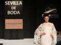 Modelo en vestido blanco con detalles encajados camina por el escenario de 'Sevilla de Boda'. En el fondo, se puede leer 'Pronovias Barcelona' y la marca de 'Feria Sevilla'.