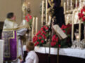 Una imagen religiosa con una figura de madera en un altar, rodeada de flores rojas y velas blancas. Un hombre vestido con un hábito religioso está bendiciendo a una niña en el altar.