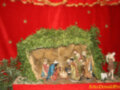 Escena navideña con un nacimiento en una cripta decorada con árboles de navidad. La escena incluye figuras de la Virgen María, el Niño Jesús y los pastores. El fondo es una cortina roja con estrellas doradas.