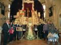 Fotografía de una familia posando junto a una imagen religiosa en un templo. La imagen muestra a varias personas, incluyendo adultos y niños, vestidos formalmente, posando frente a una estatua de la Virgen María. La escena se desarrolla en un ambiente religioso, con detalles como vitrales y una araña de luces. La familia parece estar celebrando o participando en un evento religioso importante.