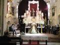 Una iglesia con un altar central, flores y velas. Un hombre en el púlpito habla a una audiencia sentada, con un altar dorado y una imagen central. La decoración es elegante y religiosa.