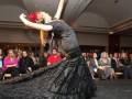 Una bailarina en un vestido negro con volantes realiza una coreografía de baile flamenco mientras un público atento observa desde la audiencia.