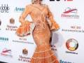 Una mujer con un vestido naranja de flamenca posa en el Salón Internacional de la Moda Flamenca 2012, con un fondo que muestra los logos de los patrocinadores.