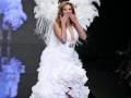 Una modelo con un vestido blanco y alas de ángel desfilando en una pasarela.