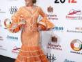 Fotografía de una mujer en un vestido de flamenca naranja brillante, posando frente a un fondo con logos de SIMOF 2012.
