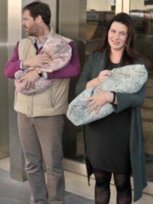 Una pareja con una persona embarazada y dos bebés recién nacidos. La imagen muestra a la mujer con un vestido largo y el hombre con una chaqueta de cuadros. El ambiente parece ser urbano, posiblemente en una estación o entrada de edificio.