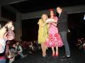Una modelo en falda de flamenco sostiene un premio mientras una mujer y un hombre la saludan. La escena se desarrolla en un escenario con público asistente observando la celebración.