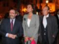 Tres personas posan juntas en una noche iluminada. La imagen muestra a dos hombres y una mujer, todos vestidos formalmente. El hombre de la izquierda lleva gafas y un corbata roja, el del medio tiene una camiseta blanca y una chaqueta gris, y el hombre de la derecha usa un traje negro con corbata azul. La escena parece ser de una celebración o evento formal, con luces y edificios en el fondo.