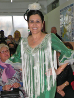 Una mujer sonríe ampliamente mientras posa para la foto. Lleva un vestido verde con detalles blancos y una flor en el cabello. La imagen se centra en ella, destacando su expresión y vestimenta.