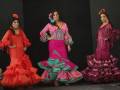 Tres modelos vestidas con trajes de flamenco posan en una pasarela. La imagen muestra trajes coloridos con volantes y adornos florales típicos de la danza española.