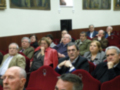 Una audiencia en un salón de conferencias, con personas sentadas en filas. La imagen muestra a varias personas de diferentes edades y estilos vestimentarios, sentadas en sillas rojas. En el fondo, se puede ver una pared blanca con un cartel y una ventana.