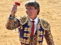 Un torero en traje tradicional sostiene una flor roja, con un fondo de arena.