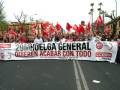 Un grupo de personas con pancartas rojas y banderas ondeando en una manifestación. En el centro, un cartel grande con la fecha 29-M y el texto "HUELGA GENERAL QUIEREN ACABAR CON TODO". La imagen muestra una calle con árboles y edificios al fondo.