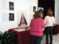 Dos personas observan una exposición de arte, con carteles y fotografías en el fondo. La imagen muestra detalles artísticos y una pieza central con un vestido de lujo.