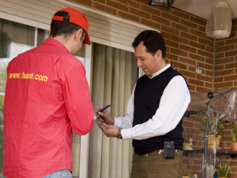 Un hombre en una chaqueta roja con el logotipo de www.huse.com está firmando un documento mientras otro hombre en una camisa blanca y un chaleco negro lo observa.