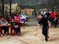 Un grupo de personas juega un juego de pelota en una zona abierta. Algunos están sentados en sillas rojas, mientras que otros se encuentran de pie. En el fondo, hay árboles sin hojas y una casa con un tejado rojo. La escena parece ser en un día soleado, posiblemente en primavera o verano.