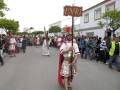 Desfile histórico con personajes de época, incluyendo un hombre vestido como soldado romano sosteniendo una bandera con la inscripción "INRI". La escena se desarrolla en un ambiente urbano, con edificios y árboles al fondo.