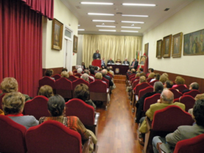 Una sala de conferencias con una larga mesa central y asientos rojos para los asistentes. La sala está iluminada con luces de techo y tiene cortinas en la parte posterior.