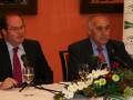 Dos hombres en trajes se encuentran frente a un microfono, con una mesa de cristal y flores en el centro. En el fondo, hay un cartel con logotipos y texto que parece ser de una conferencia o evento.
