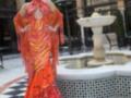 Una mujer vestida con un vestido de flamenca color naranja y rojo, posando frente a una fuente en un patio con baldosas negras y blancas.