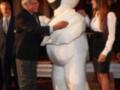 Un hombre en traje abraza a un personaje inflable de un pingüino de dibujos animados en una alfombra roja. Una mujer observa la escena.